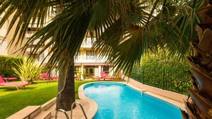 Une piscine avec des marches permettant d’y accéder, entourée de palmiers et, au fond, un bâtiment doté de balcons.