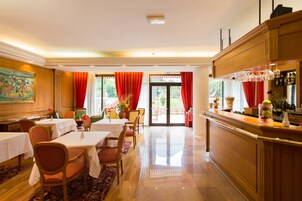 A hotel dining area with tables and chairs, a bar with glasses and bottles, and a view of the outdoors through large windows.