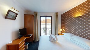 A hotel room with a bed, a desk with a television, a picture frame, a lamp, and a view of the outside through a window. - Sun Riviera (Cannes)