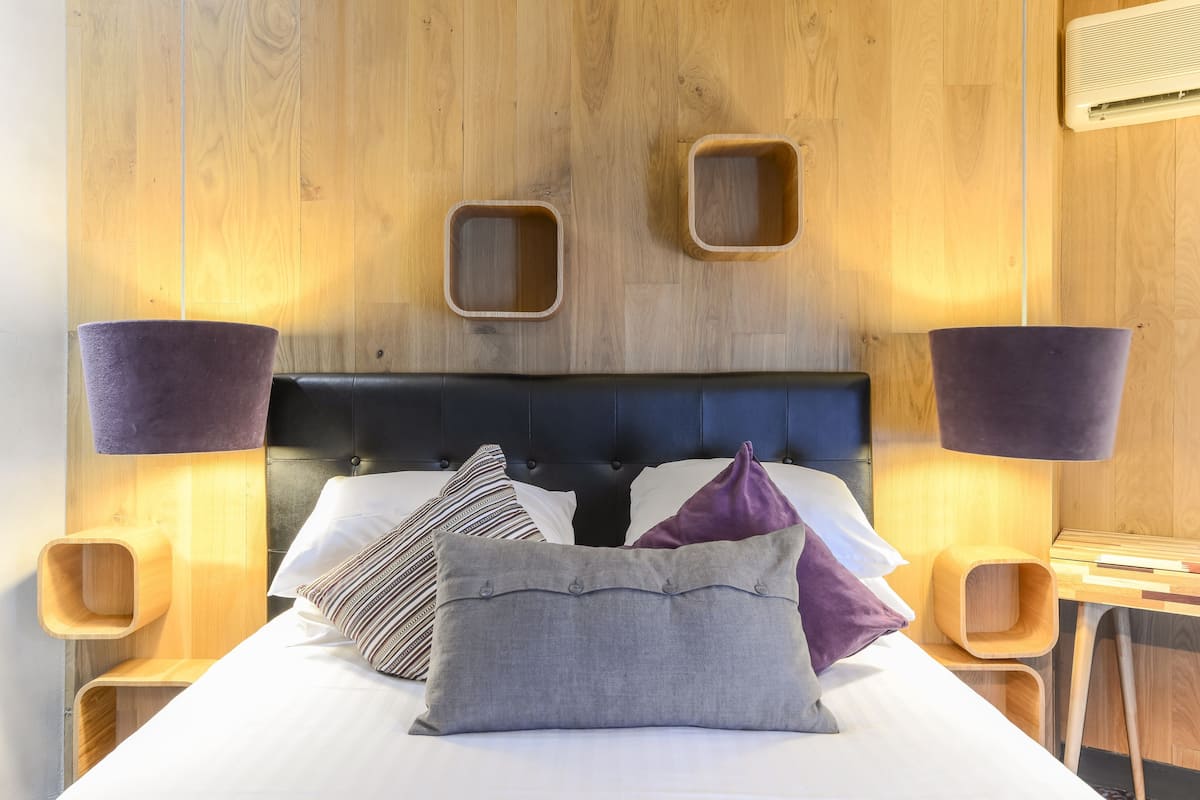 A neatly made bed with a dark headboard, two bedside tables with lamps, and a wooden wall with built-in shelves.