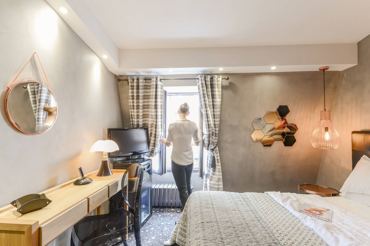 A hotel room with a bed, a desk with a lamp, a TV, and a view of the outside through curtains.