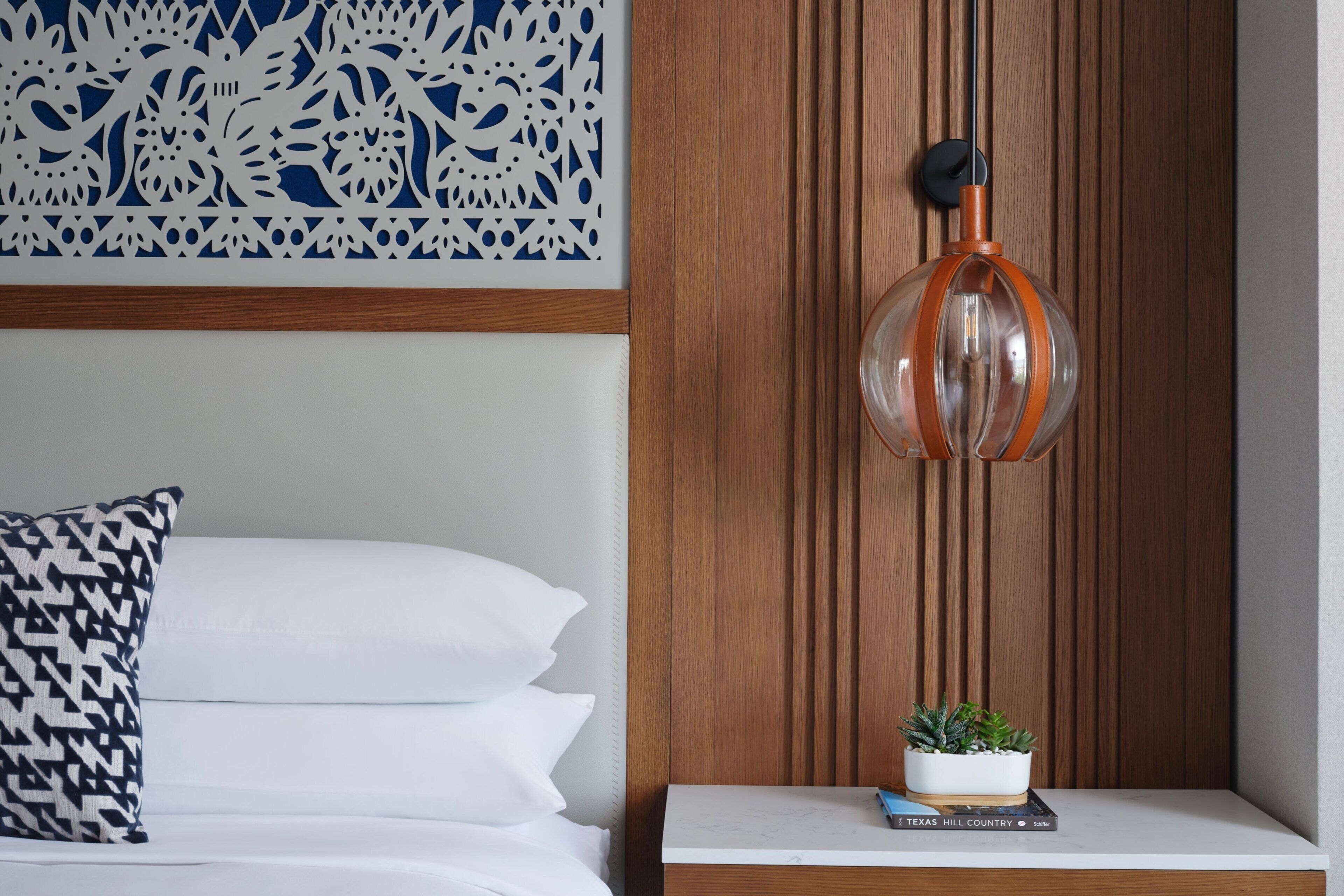 A neatly made bed with white and black pillows, a decorative wall panel, a clear glass pendant light, and a small plant on a white nightstand.
