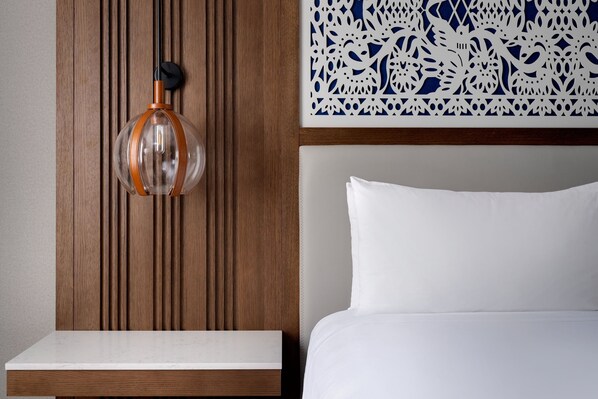Habitación de hotel con pared de paneles de madera, un tapiz decorativo, cama con ropa blanca y una lámpara colgante de cristal transparente con detalle naranja.