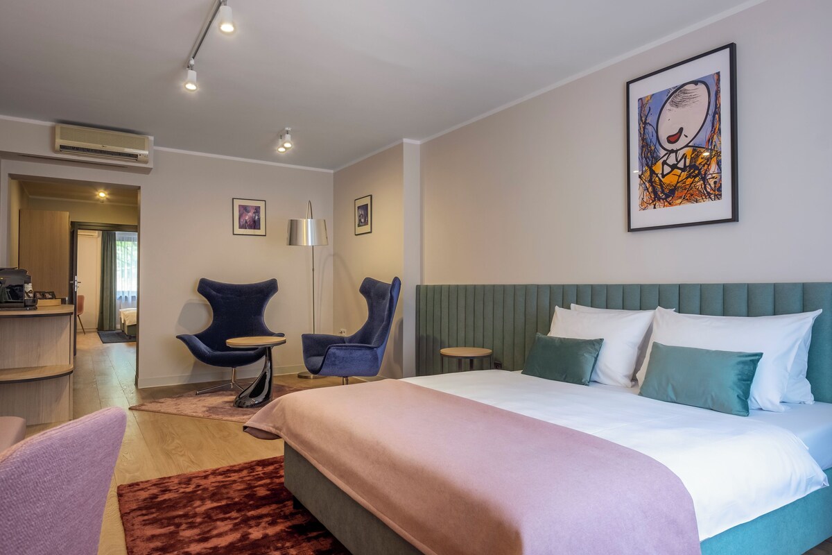 A hotel room with a bed, two chairs, a desk, and a framed artwork on the wall.