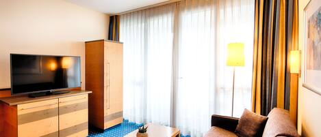 A hotel room with a flat-screen TV, a wooden cabinet, a sofa, a small table, and a large window with curtains.
