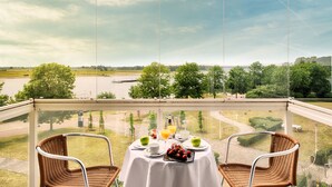 A balcony with two wicker chairs, a table with breakfast items, and a scenic view of a river and trees. - Welcome Hotel Wesel (Wesel)