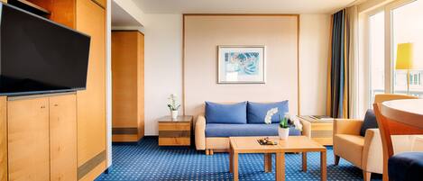 Habitación de hotel con alfombra azul estampada, un sofá, una mesa de centro, un televisor y una ventana con cortinas.