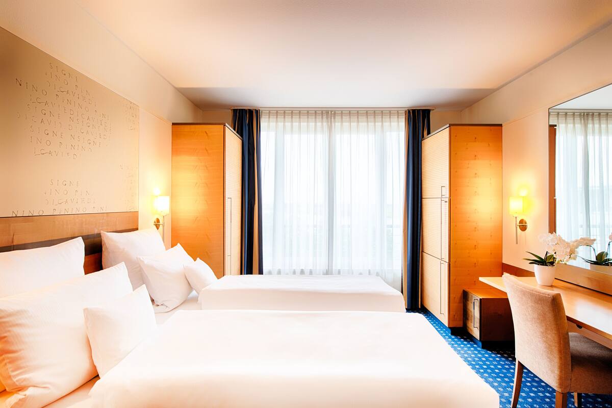 A hotel room with a large bed, a desk, and a chair. There is a window with curtains, a wall with writing, and a lamp on the desk.