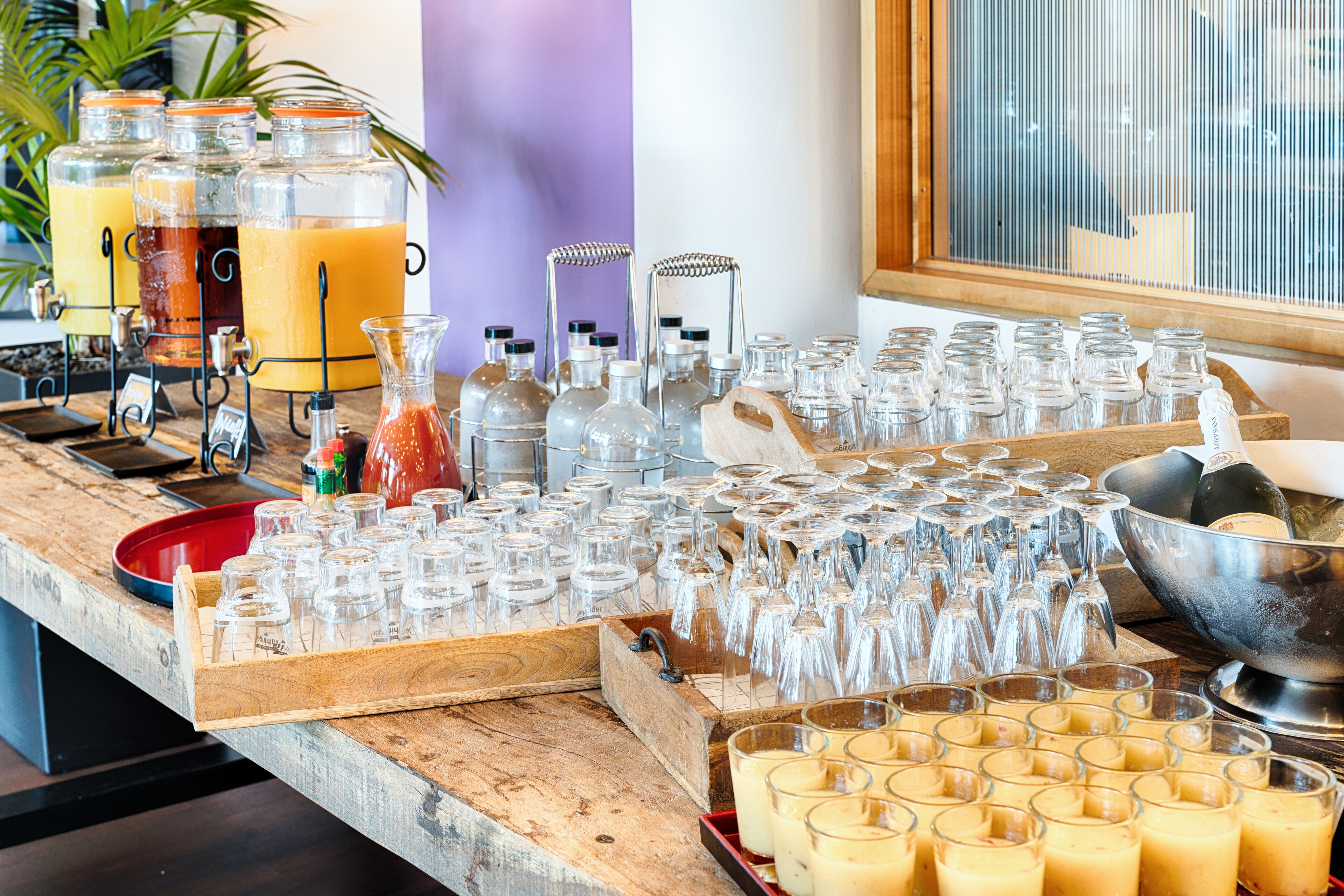 A buffet setup with various drink dispensers, a large number of glasses, and a wooden serving tray.