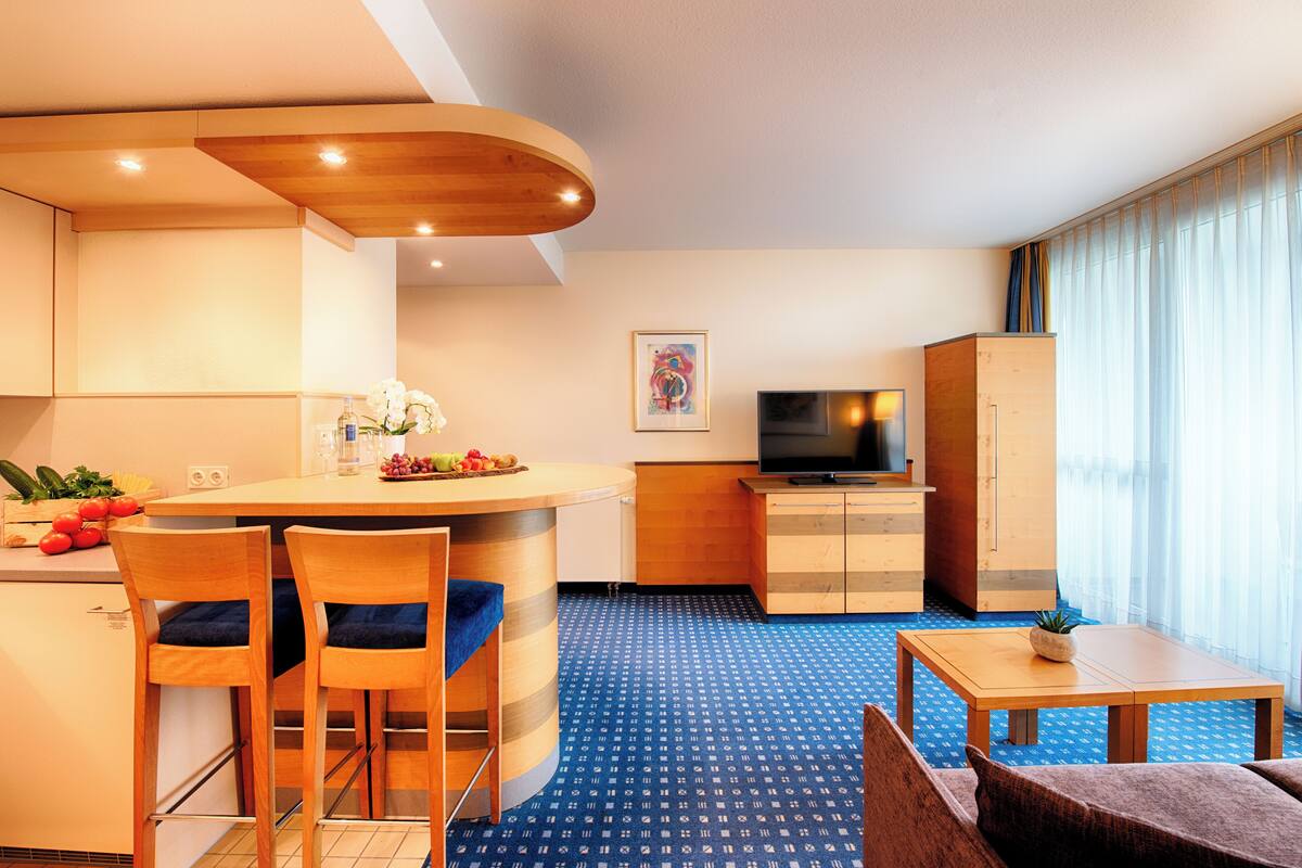 A compact hotel room with a kitchenette, a dining area with two chairs, a small TV, and a large window with curtains.