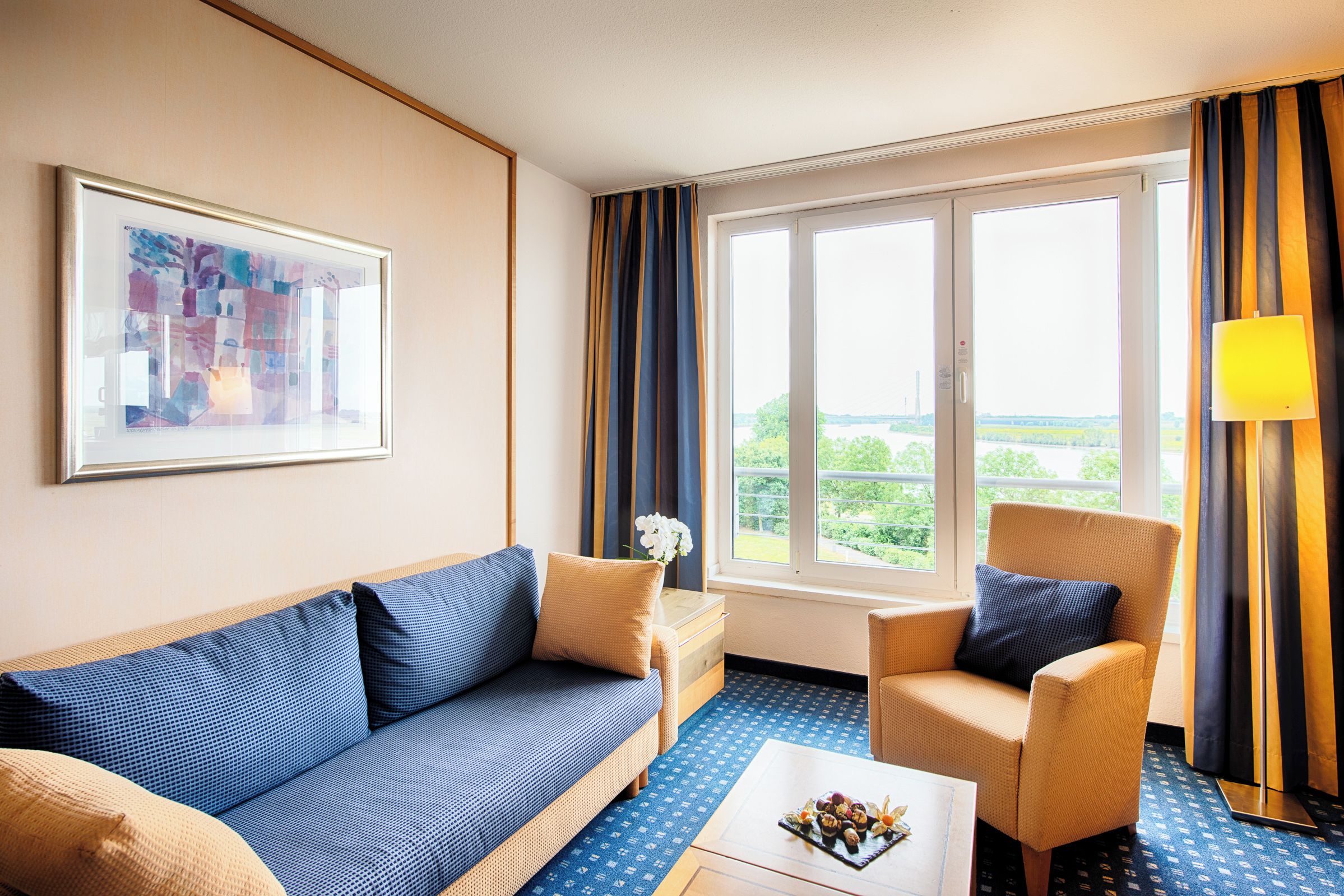 Habitación de hotel con sofá, sillón y vistas a zonas verdes por la ventana.