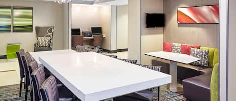 A conference room with a large white table, black chairs, a sofa with colorful cushions, and a TV on the wall.