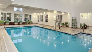 An indoor swimming pool with clear water, surrounded by green chairs and tables, and large windows allowing natural light.