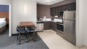 A compact hotel room with a kitchenette, a bed with white linens, a small dining table with chairs, and a blue carpet.