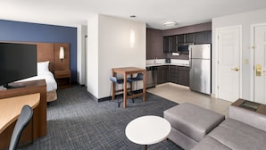 A compact hotel room with a bed, a desk, a small dining table, a sofa, a kitchen area with a refrigerator and microwave, and a TV.