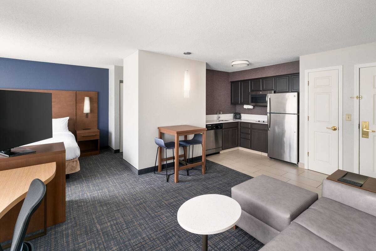 A compact hotel room with a bed, a desk, a small dining table, a sofa, a kitchen area with a refrigerator and microwave, and a TV.
