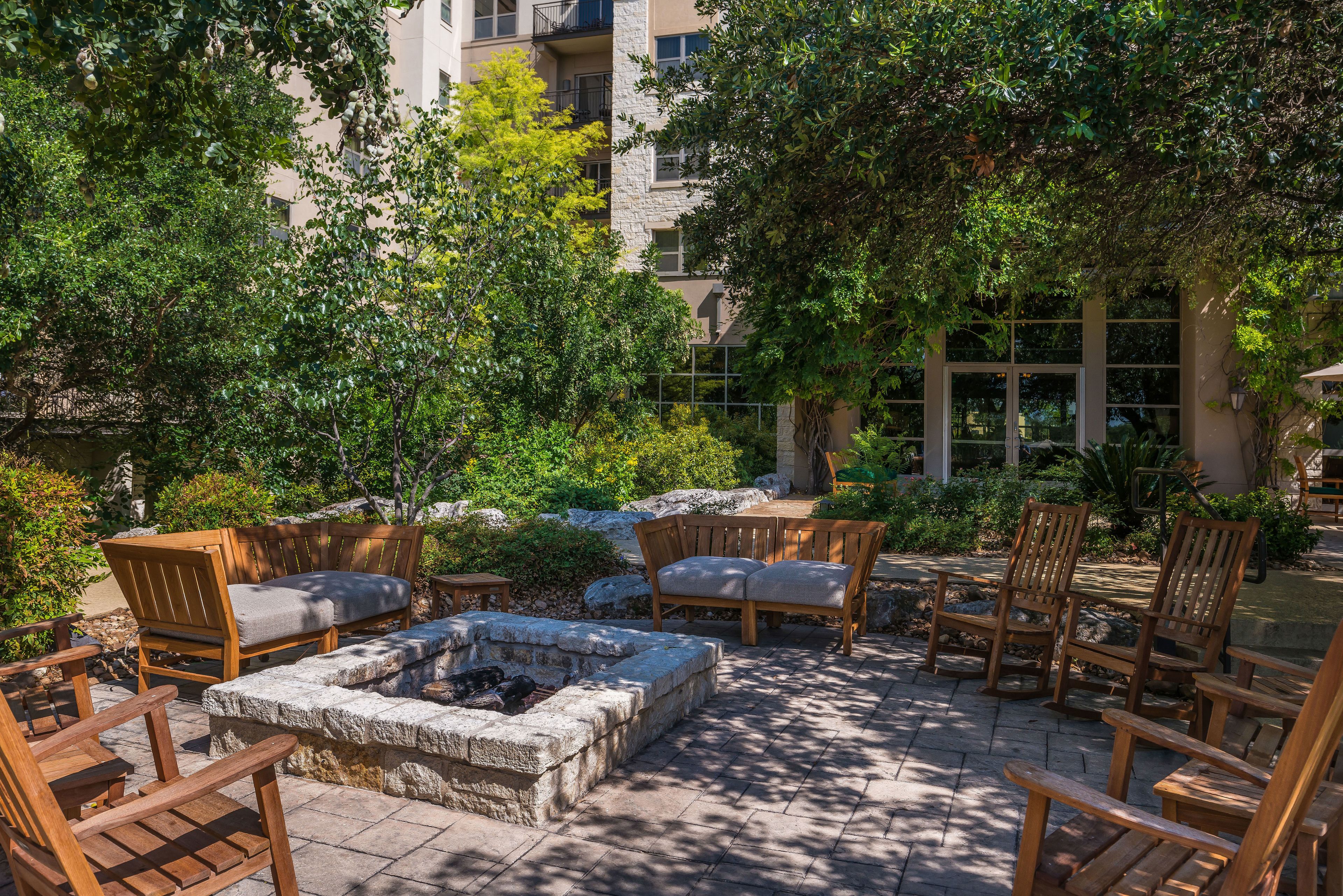 Un patio al aire libre con sillas de madera y un hogar de piedra rodeado de abundante vegetación y un edificio de piedra al fondo.