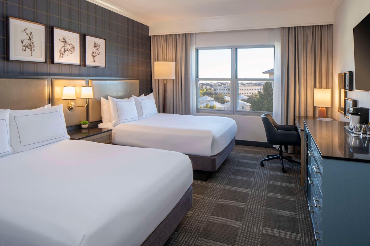 A hotel room with two beds, a desk, a chair, and a large window with a city view.