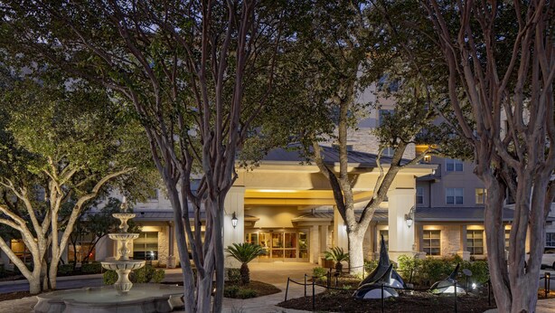 A hotel entrance with a fountain, trees, and a well-lit driveway.