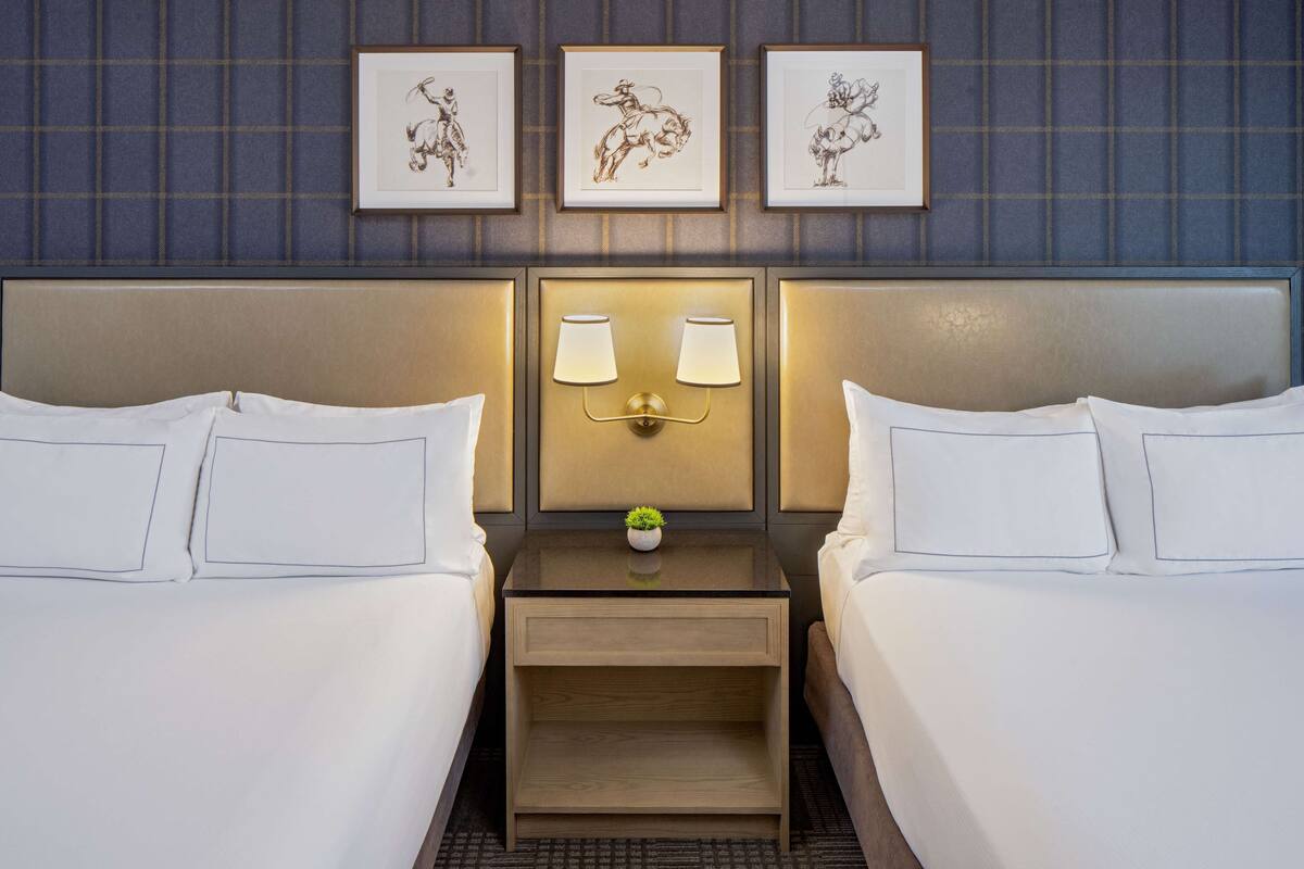 A hotel room with two beds, a nightstand with lamps, and three framed artworks on the wall.