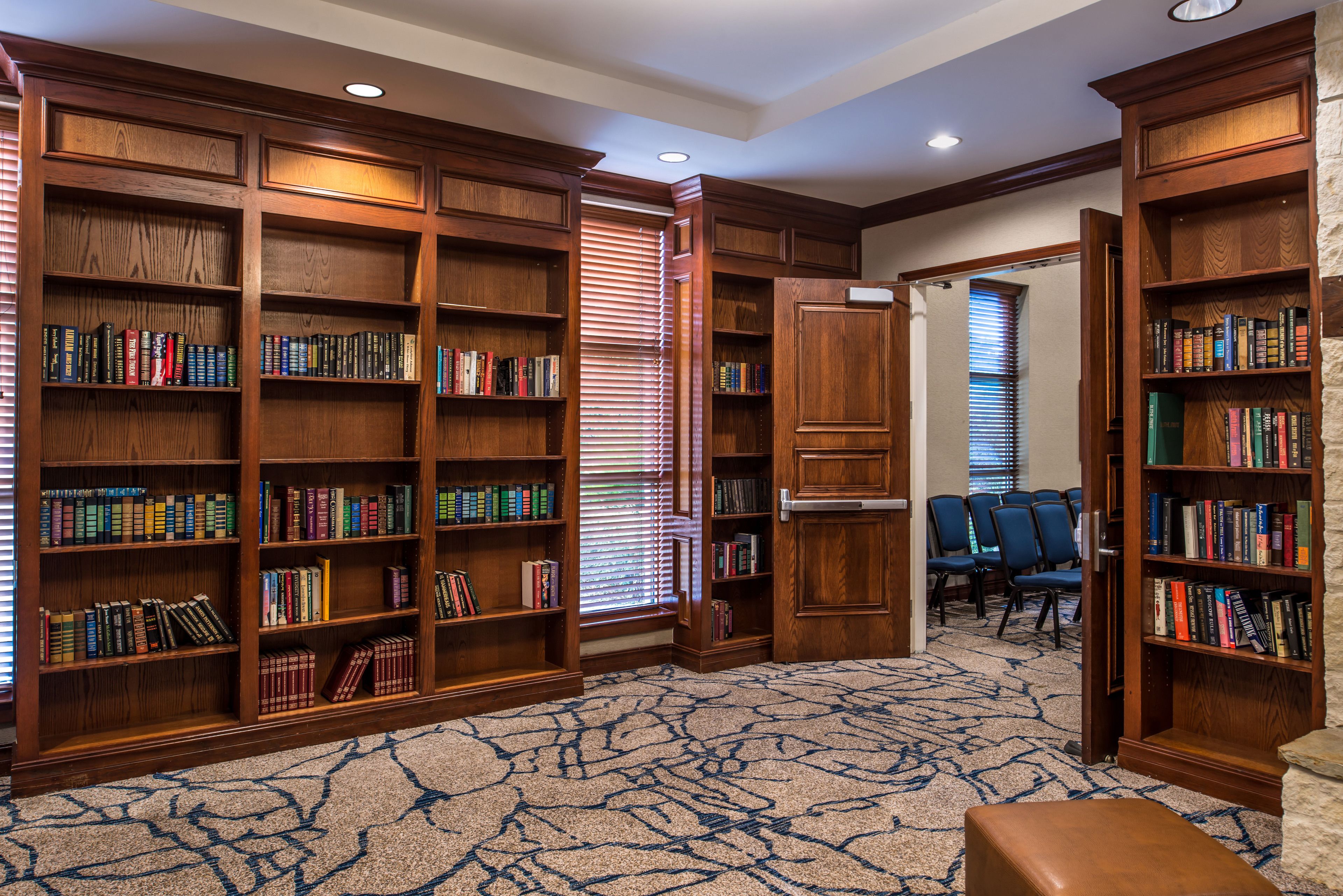 A library with wooden bookshelves, a patterned carpet, and a seating area with chairs.