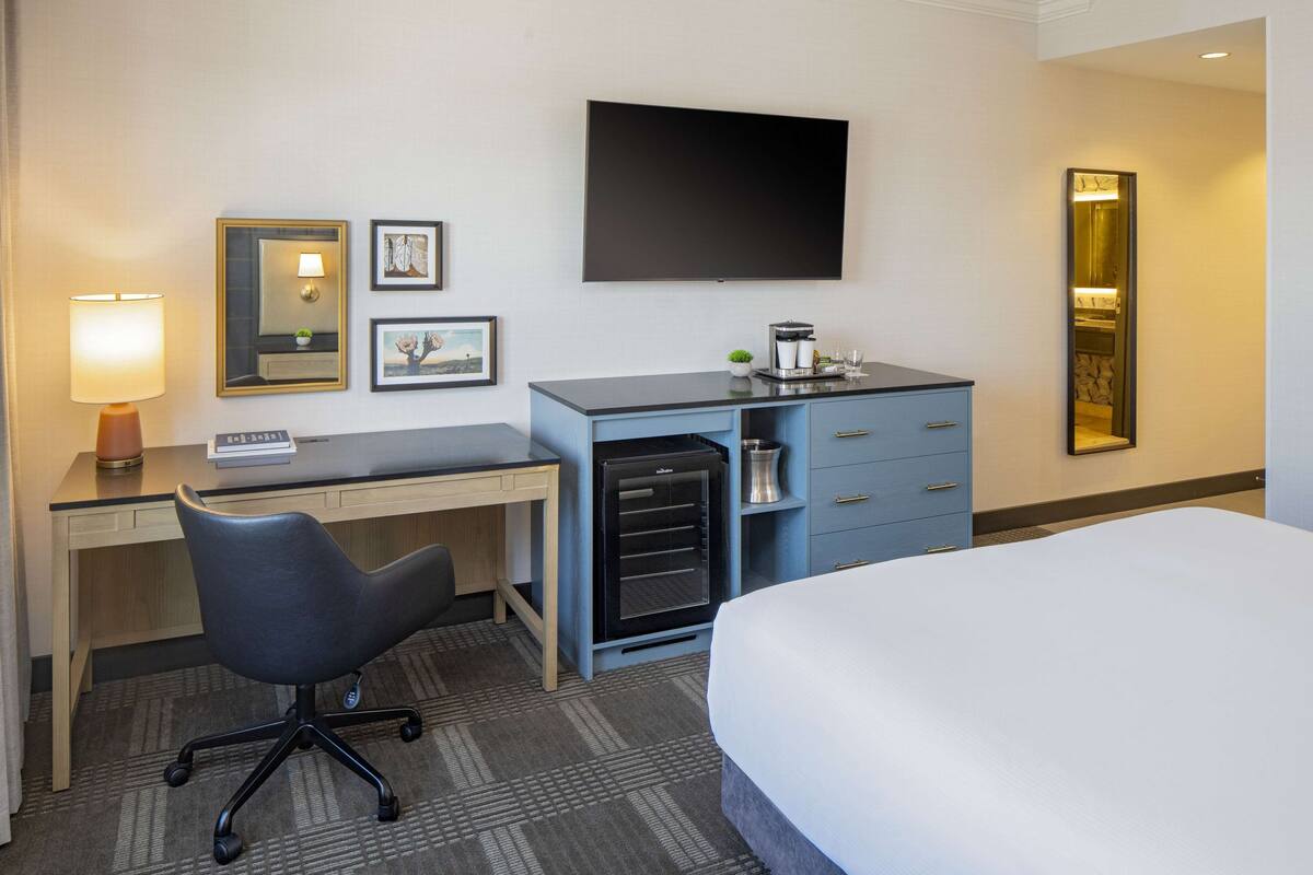 A hotel room with a bed, desk, chair, and a flat-screen TV mounted on the wall.