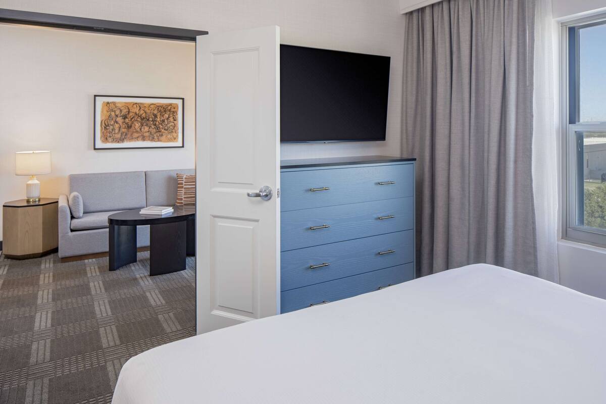 A hotel room with a bed, a blue dresser, a television, a sofa, a lamp, and a painting on the wall.