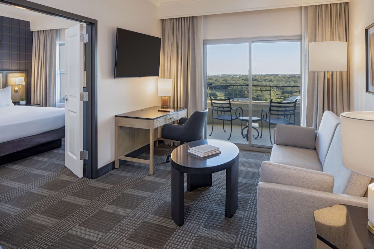 A hotel room with a bed, desk, chairs, a small table, and a balcony with a view.