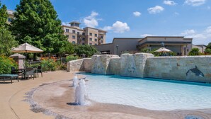 A swimming pool with a water feature, surrounded by lounge chairs and umbrellas, with buildings in the background.