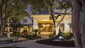 A hotel entrance with a fountain, trees, and a well-lit pathway leading to the entrance.