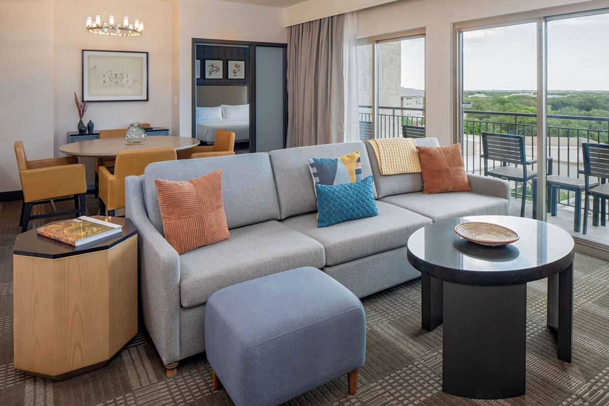 A modern hotel room with a sofa, ottoman, dining table, and chairs. There is a balcony with a view of the city.