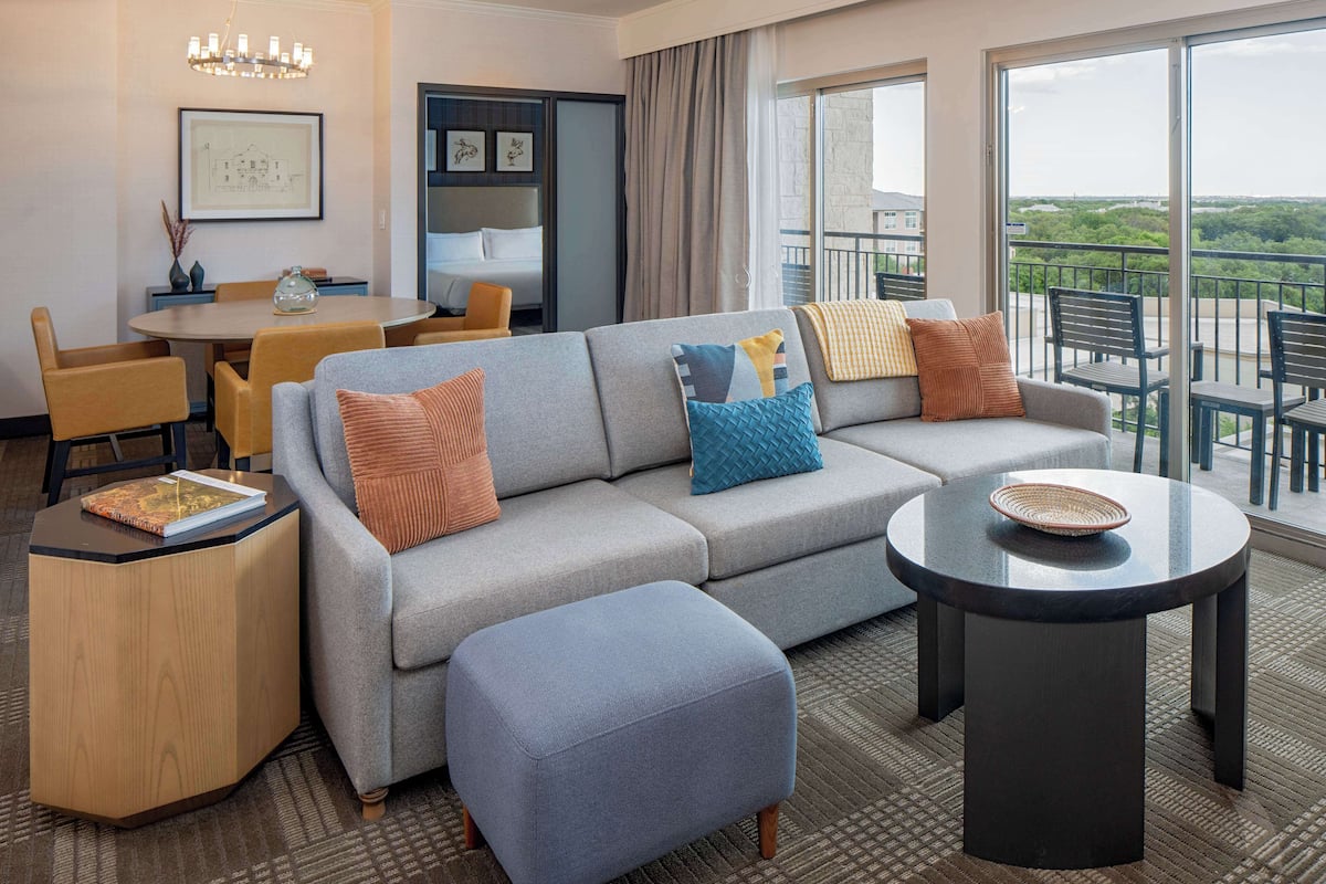 A modern hotel room with a sofa, ottoman, dining table, and chairs. There is a balcony with a view of the city.