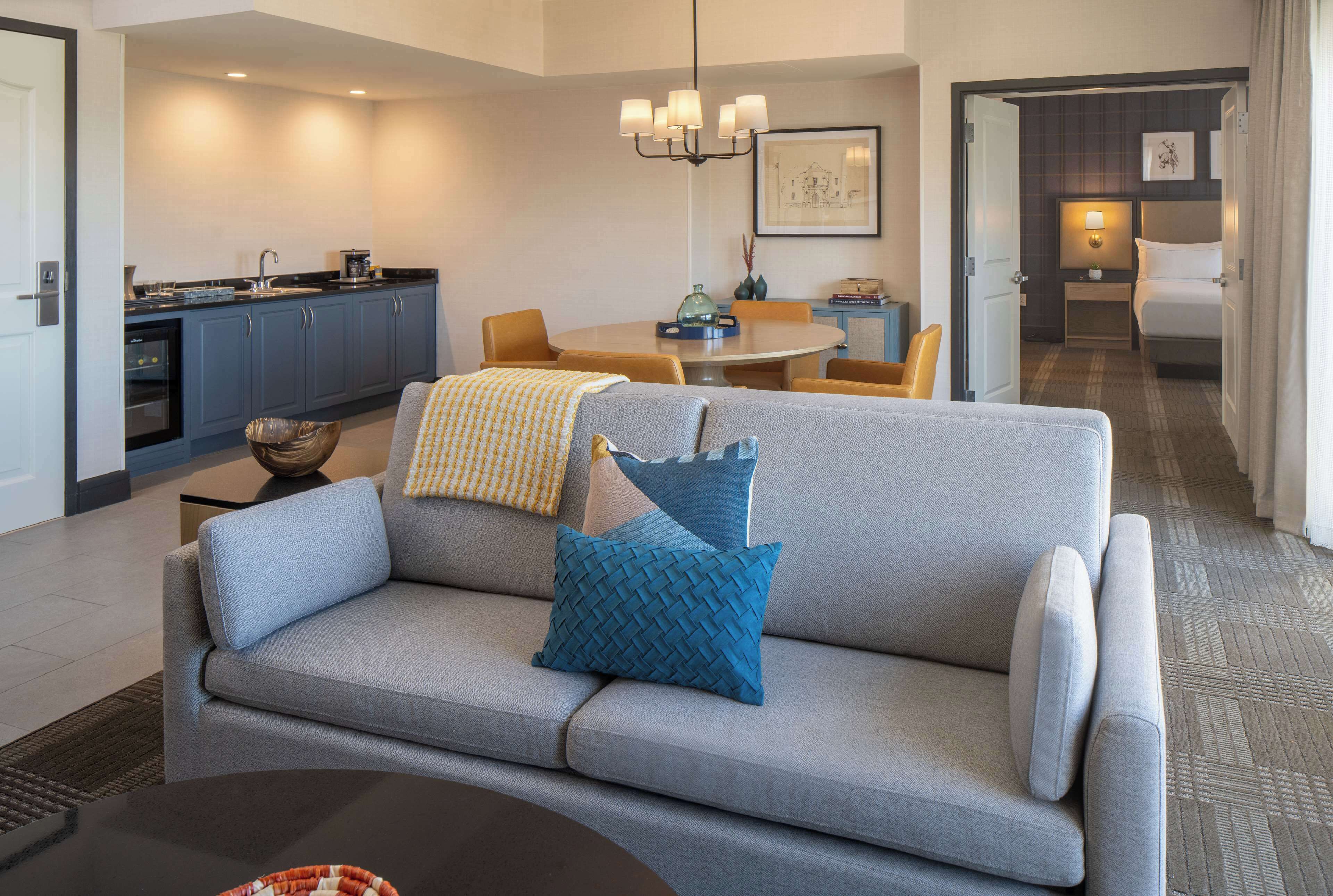 Ein modernes Hotelzimmer mit grauem Sofa, einem Esstisch, einer Küchenzeile und einem separaten Schlafbereich mit Bett und Nachttisch.