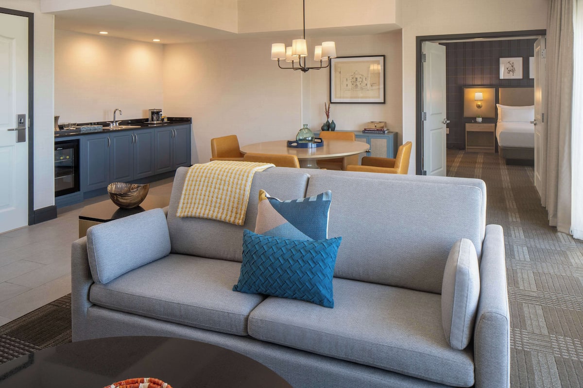 A modern hotel room with a gray sofa, a dining table, a kitchen area, and a separate bedroom with a bed and nightstand.