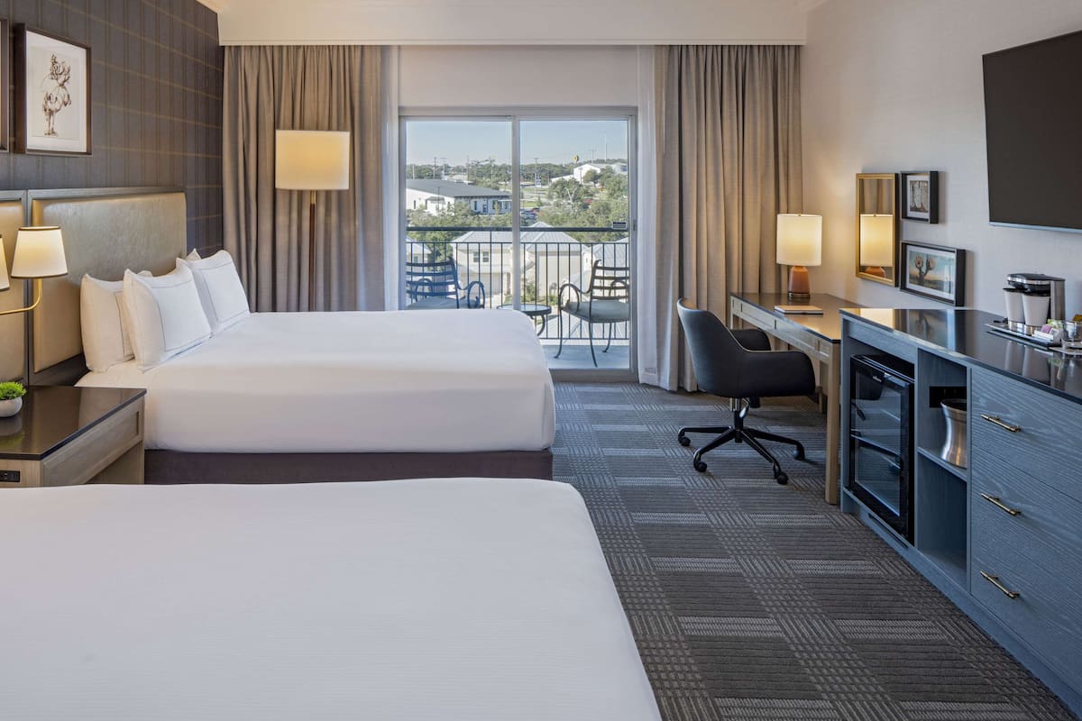 A hotel room with two beds, a desk, a chair, a TV, and a balcony with a view.