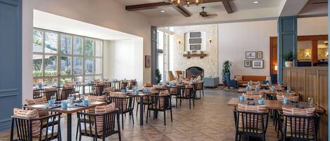 A spacious dining area with multiple tables and chairs, a stone fireplace, and large windows providing natural light.