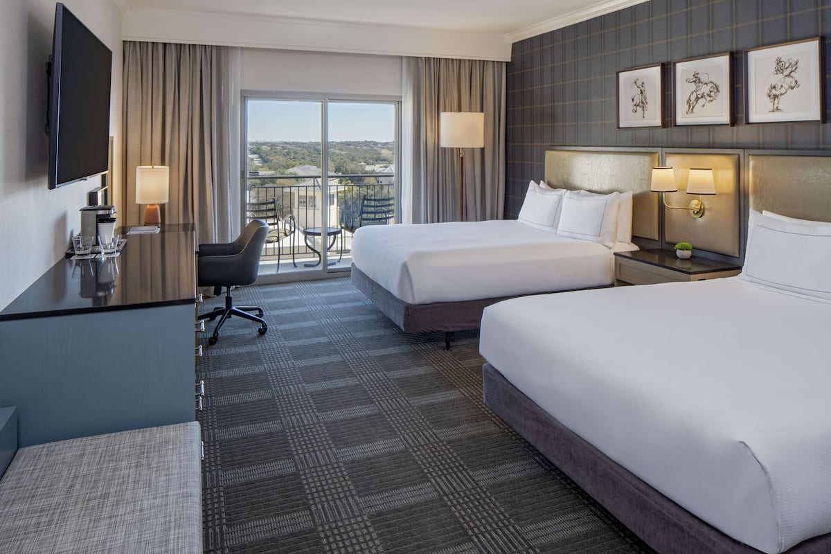 A hotel room with two beds, a desk, a chair, a TV, and a balcony with a view.