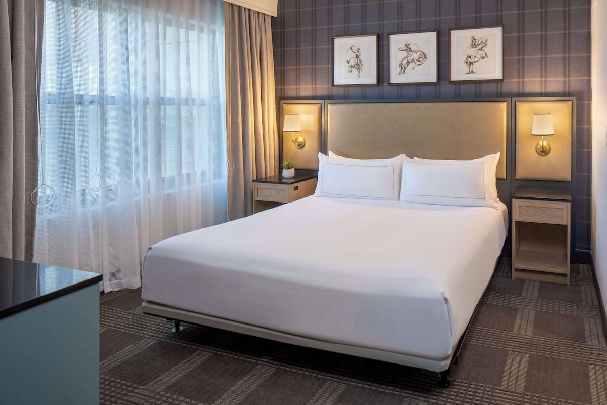 A hotel room with a large bed, two bedside tables with lamps, a desk, and a window with sheer curtains.