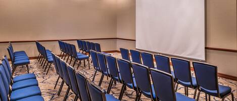 A conference room with rows of blue chairs, a carpeted floor, and a projection screen.