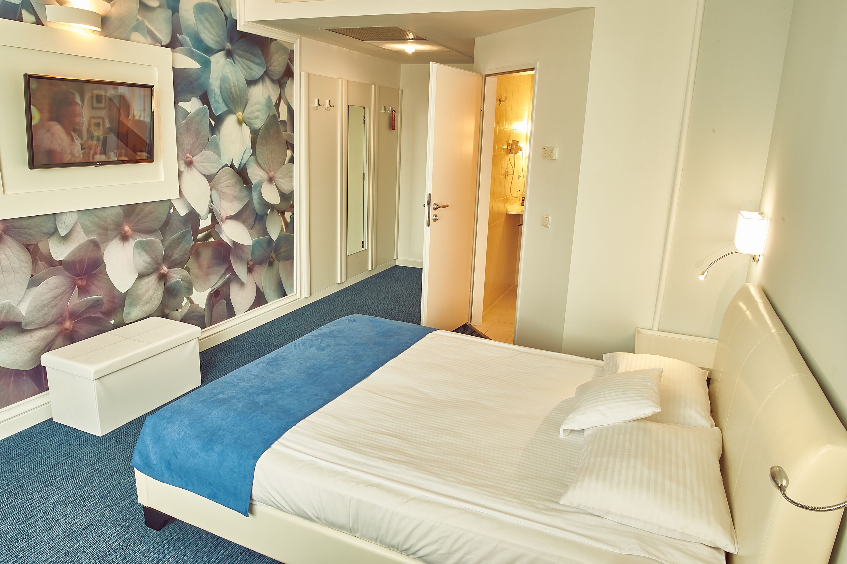 A hotel room with a bed, a bedside table, a mirror, a wall-mounted TV, and a door leading to a bathroom.