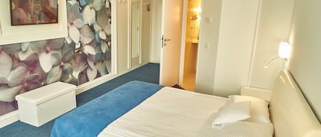 Una habitación de hotel con cama, mesita de noche, espejo, televisor montado en la pared y puerta que da a un baño.