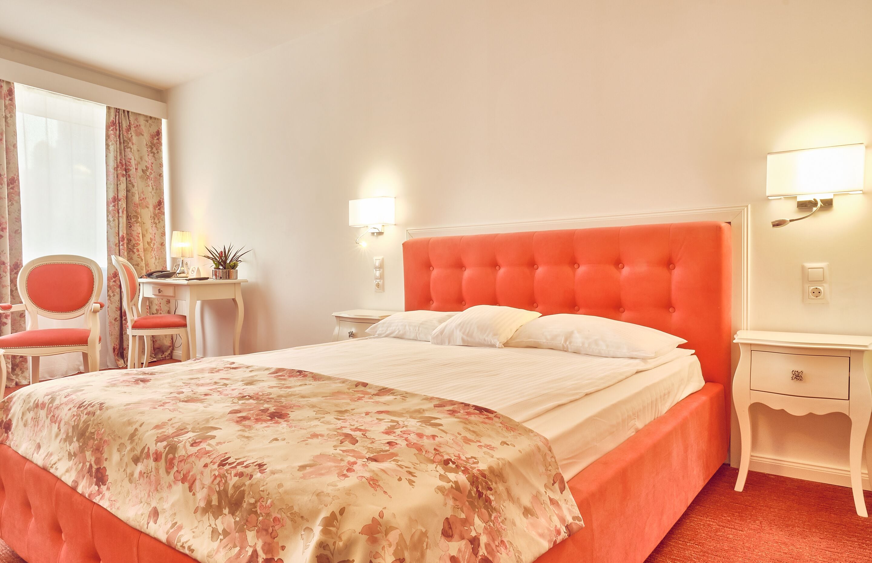 A hotel room with a large bed, a desk with a chair, a nightstand, and a window with curtains.