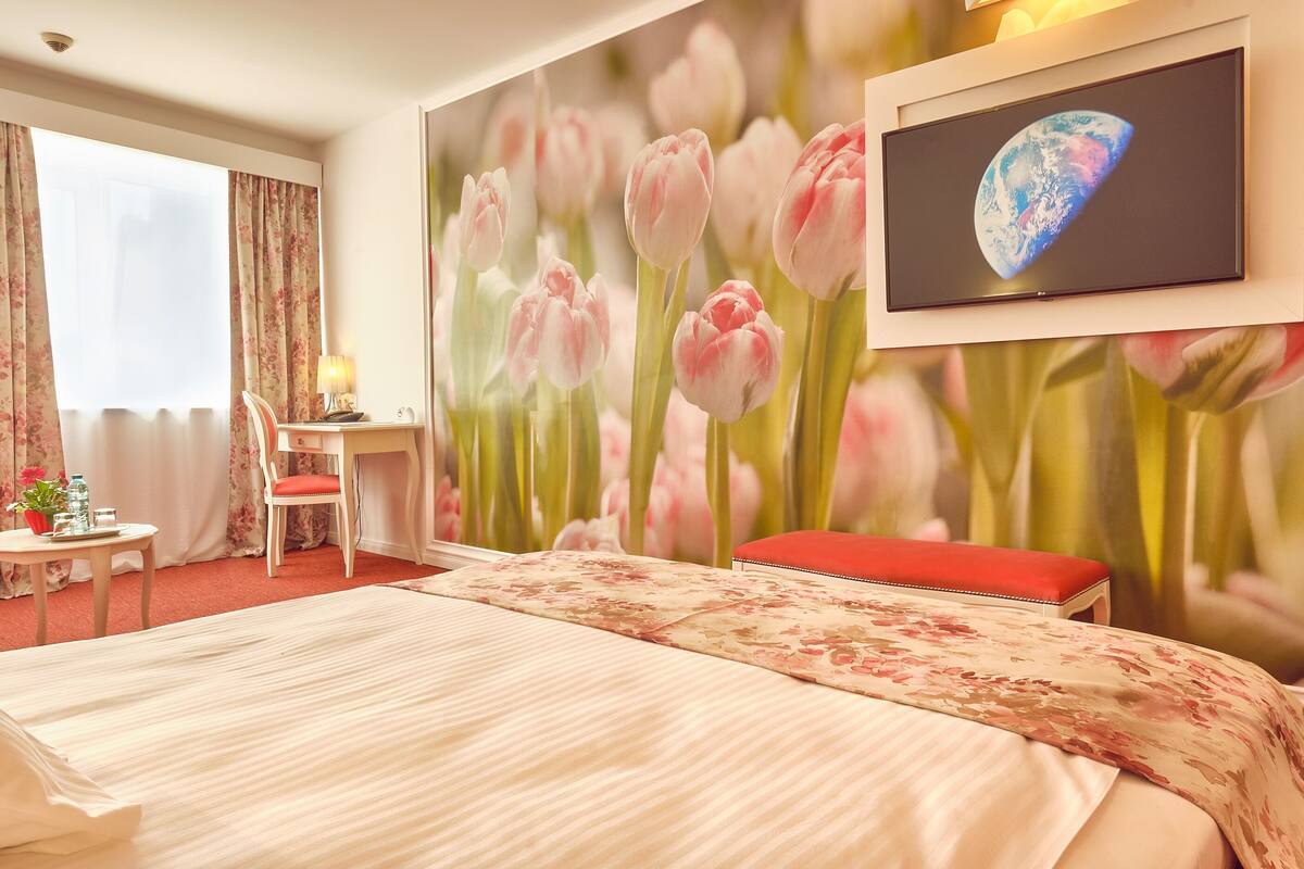 A hotel room with a large bed, a desk with a chair, a TV on the wall, and a window with curtains.