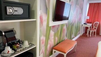 A hotel room with a coffee station, a TV mounted on the wall, a small table with a chair, and a red curtain.