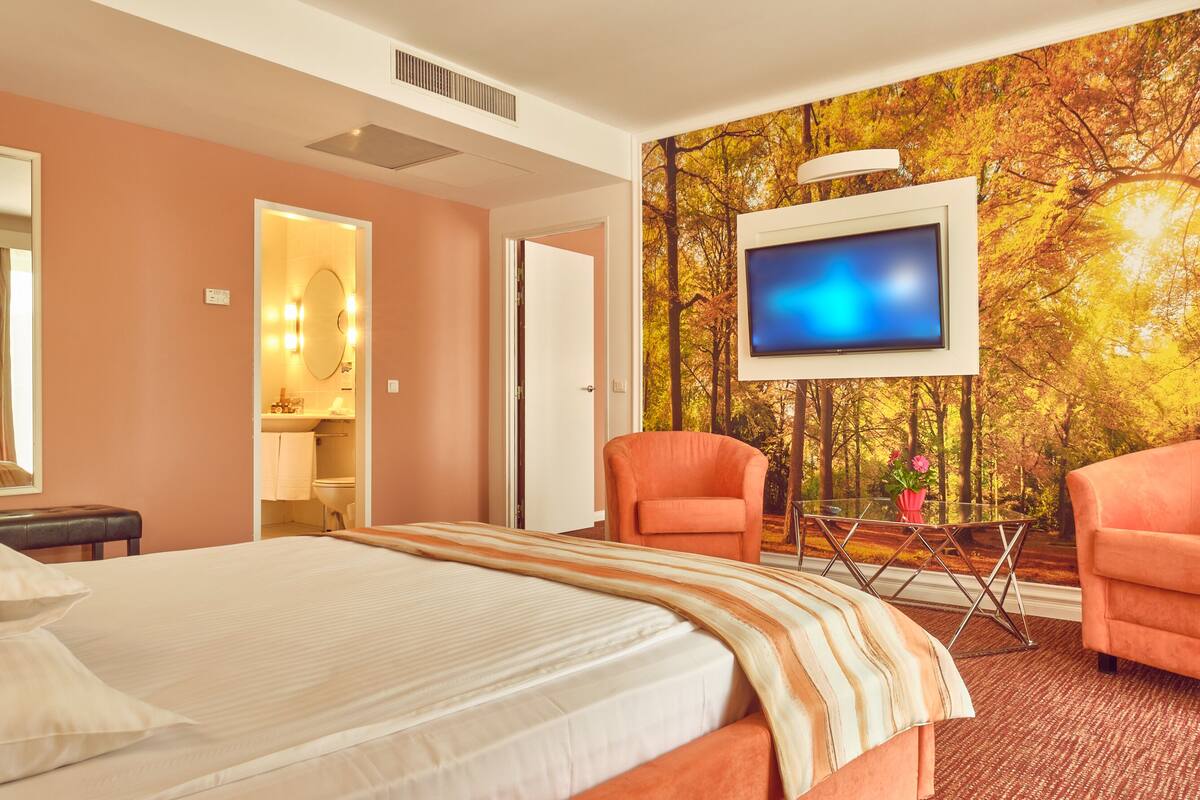 A hotel room with a bed, two armchairs, a TV, and a forest-themed wall mural.