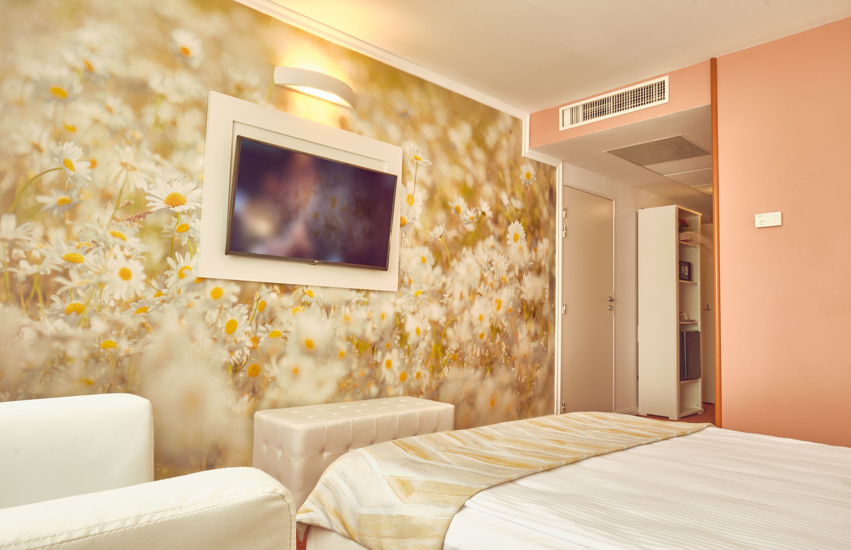 A hotel room with a floral wallpaper, a bed, a white armchair, and a television mounted on the wall.