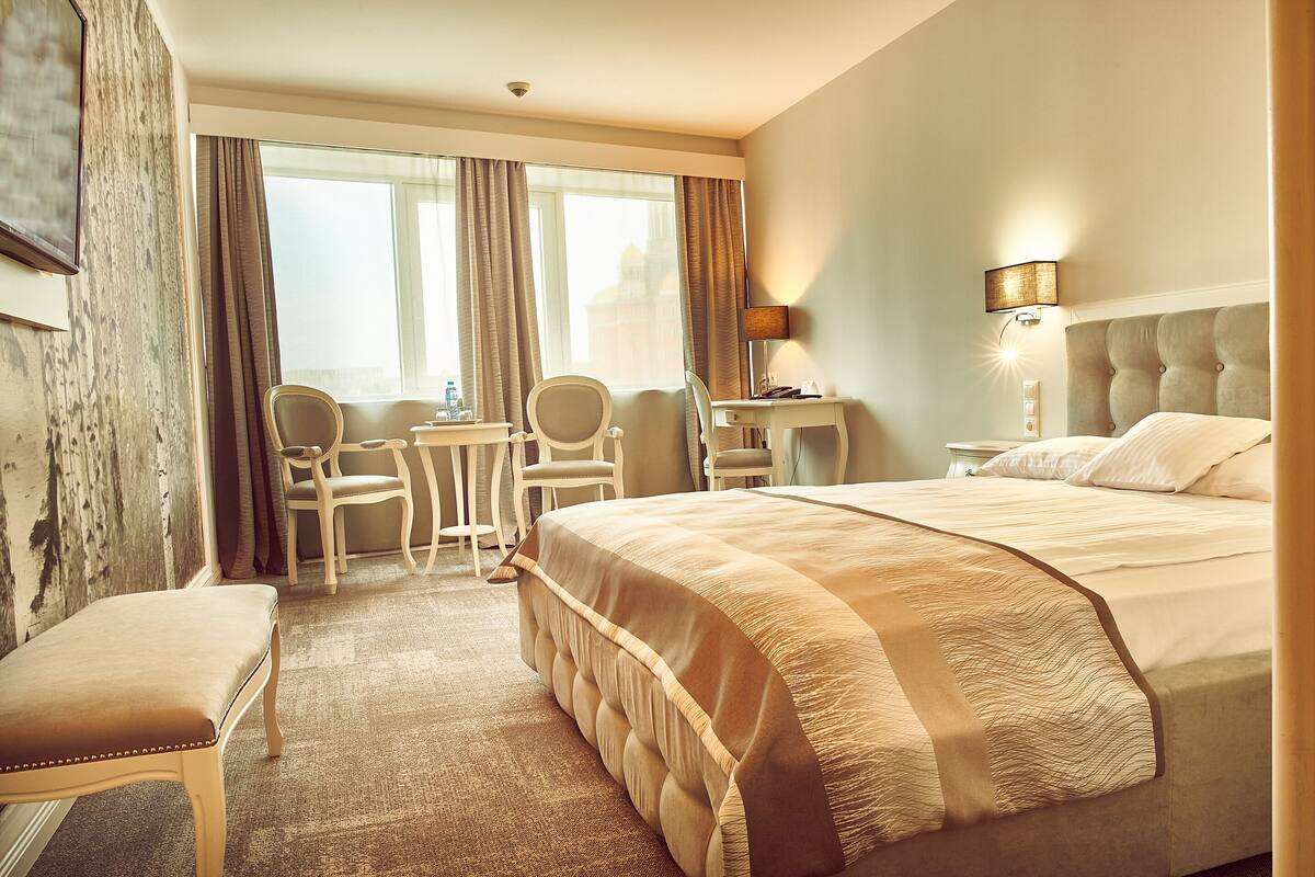 A hotel room with a large bed, a desk, two chairs, a small table, and a view of the city through the window.