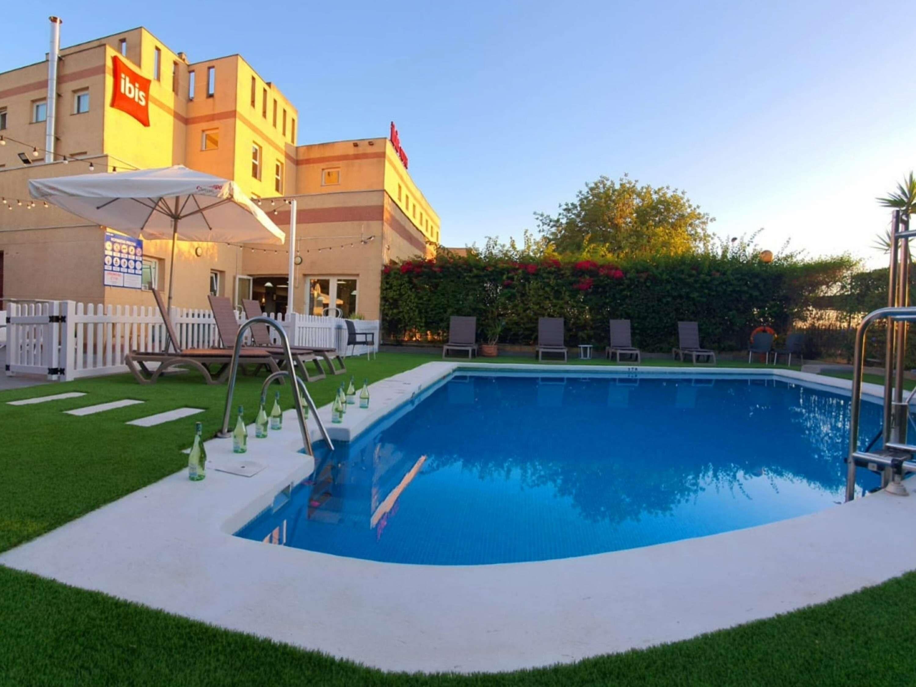 Une piscine avec des chaises longues, une clôture blanche et un bâtiment portant une enseigne « ibis ».
