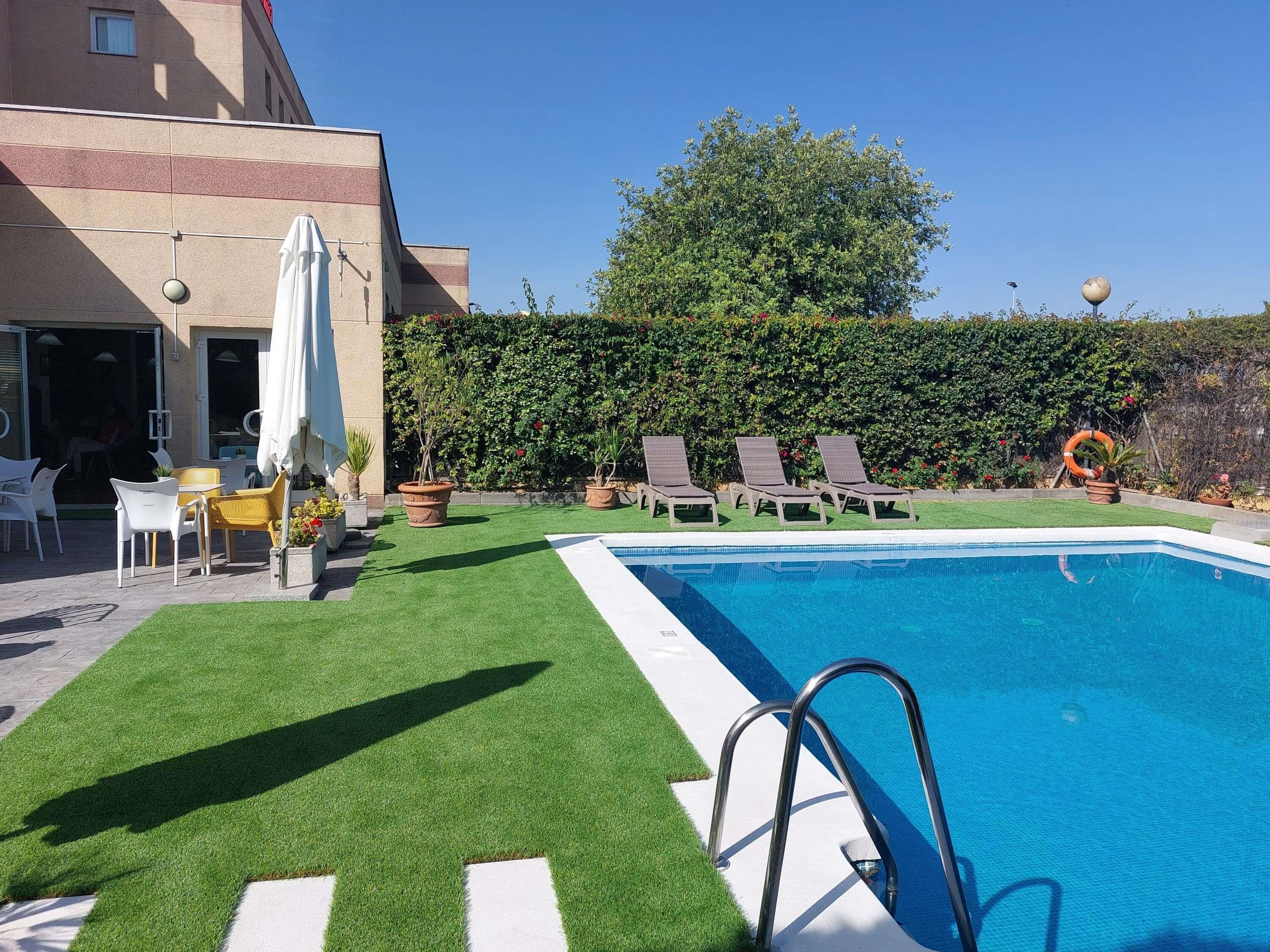 En swimmingpool med en metalstige, omgivet af kunstgræs, hvide liggestole og krukker med planter.
