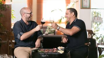 Deux hommes trinquent avec des verres de vin autour d’une table où se trouvent un gril et différentes sortes de viande.